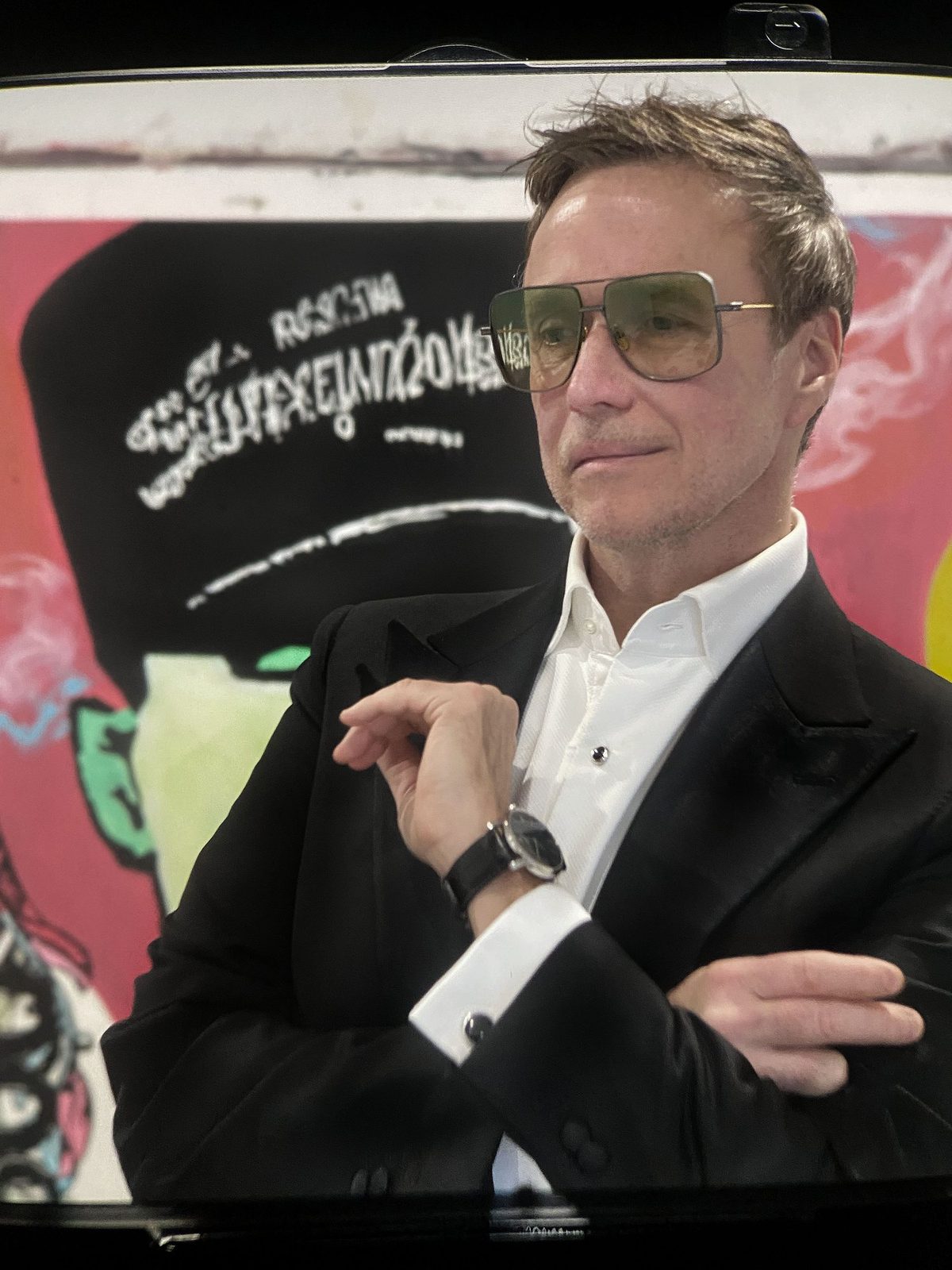 Brian Drake in a black dinner jacket and tinted aviators, arms crossed, in front of one of his paintings