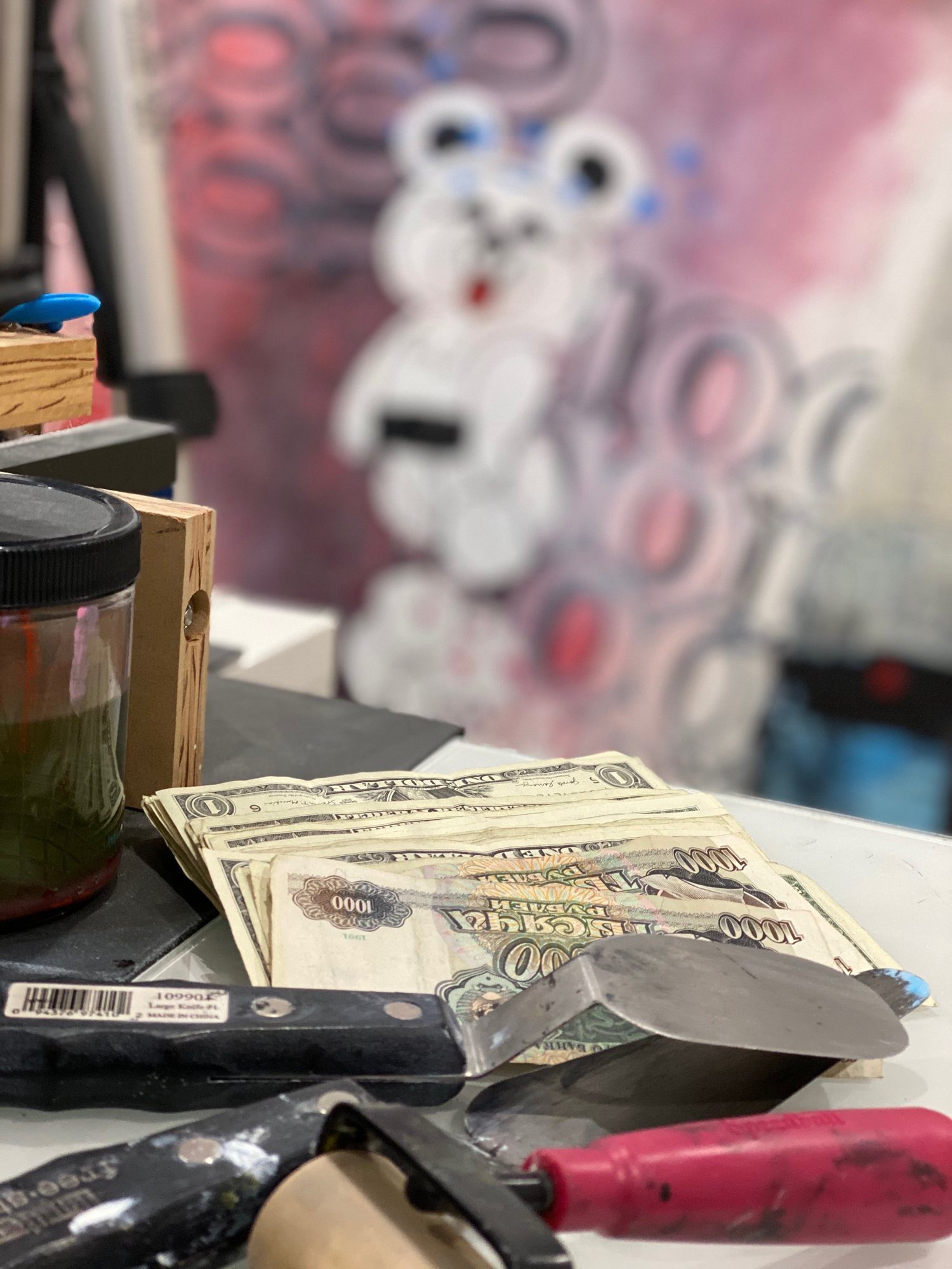 A stack of 1991 thousand-ruble notes and dollar bills beside a palette knife on the studio worktable, with a painting behind